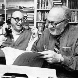 Ellery Queen photo. Black-and-white photo of two men sitting at a desk, one on the phone and one holding an open book