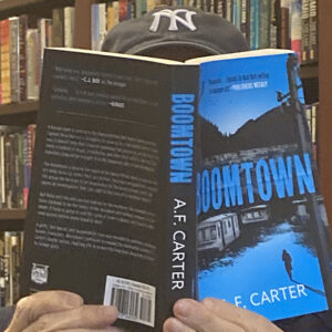 AF Carter author photo. The author's face is hidden behind a copy of Boomtown, with only a Yankees cap, blue jacket, and green shirt visible.