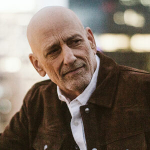 Andrew Klavan author photo. Headshot of bald man in brown jacket against a blurred background.