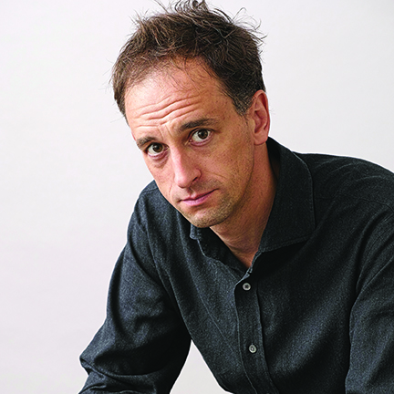 Charles Cumming author photo. Headshot of man in dark gray shirt with short, brown hair.