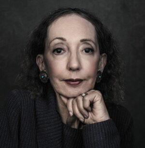 Joyce Carol Oates author photo. Headshot of woman with dark curly hair resting her chin on her fist