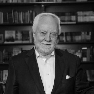 Otto Penzler author photo. Black and white image of white-haired man in a dark suit with bookshelves in the background