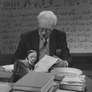 Vincent Starrett author photo. Black-and-white photo of white-haired man sitting at a desk, looking down at a book on a cluttered desk. In front of the book is a small bust of Edgar Allan Poe.