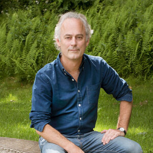 Amor Towles author photo. Color photo of a white-haired man in a blue shirt sitting outdoors.