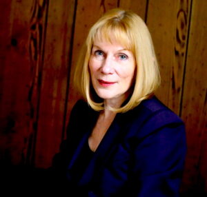 Katherine Ramsland author photo. Head shot of woman with blonde hair and bangs, wearing a blue suit.